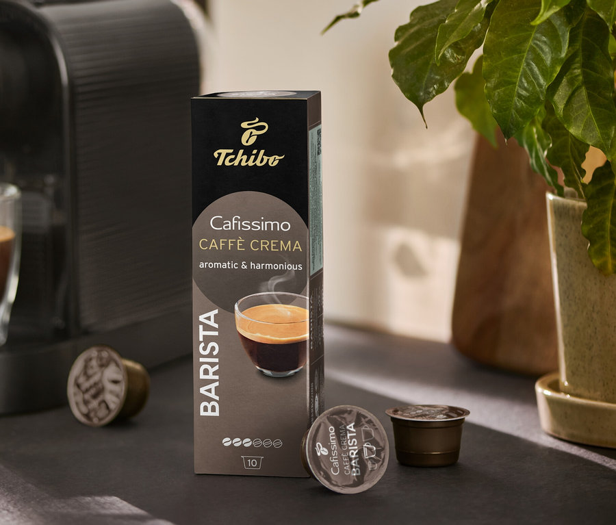 Une machine à café verse du café dans un verre. À côté se trouvent un paquet de Cafissimo BARISTA Caffè Crema et quelques capsules.