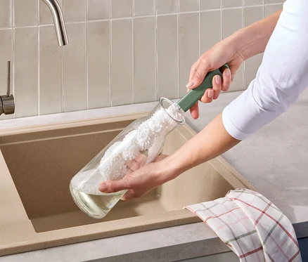 Une personne nettoie une bouteille en verre avec une brosse à bouteille verte au-dessus d'un évier beige. Un torchon à carreaux est posé sur le bord de l'évier.