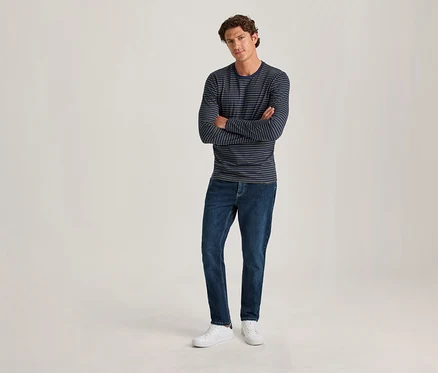 Un homme aux cheveux bruns bouclés se tient les bras croisés. Il porte un t-shirt à manches longues rayé bleu marine et bleu clair, un jean bleu foncé et des baskets blanches, sur un fond clair uni.