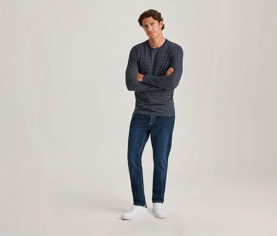 Un homme aux cheveux bruns bouclés se tient les bras croisés. Il porte un t-shirt à manches longues rayé bleu marine et bleu clair, un jean bleu foncé et des baskets blanches, sur un fond clair uni.