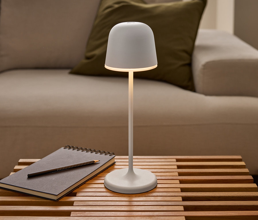 Une lampe de table à LED rechargeable Eglo « Mannera » blanche et allumée est posée sur une table en bois à lattes, à côté d'un carnet et d'un crayon, devant un canapé.