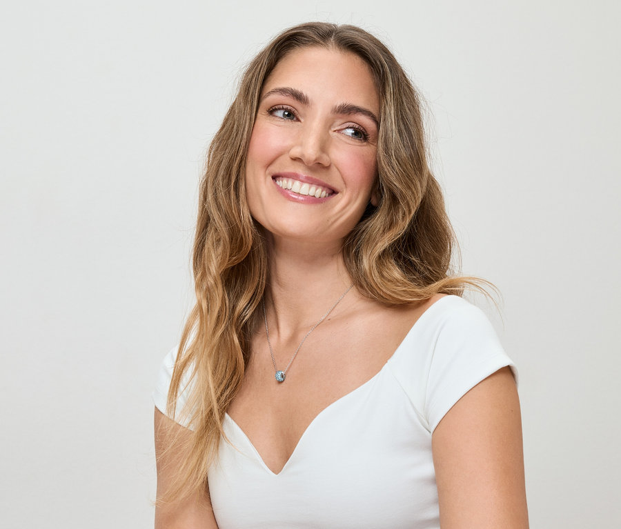 Une femme souriante aux cheveux bruns ondulés et portant un haut blanc porte un collier argenté avec un pendentif bleu.