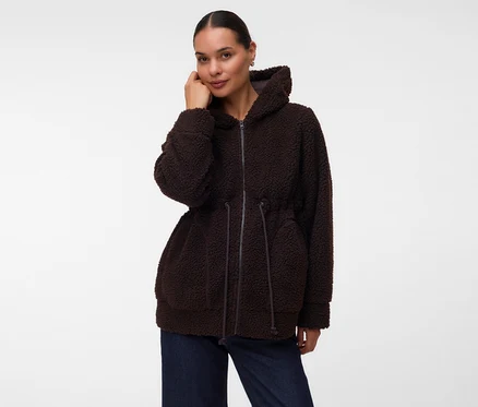 Femme portant une veste à capuche marron foncé et moelleuse avec une fermeture éclair et un cordon de serrage à la taille, associée à un jean bleu foncé. Elle regarde l'appareil photo, une main près de son visage.