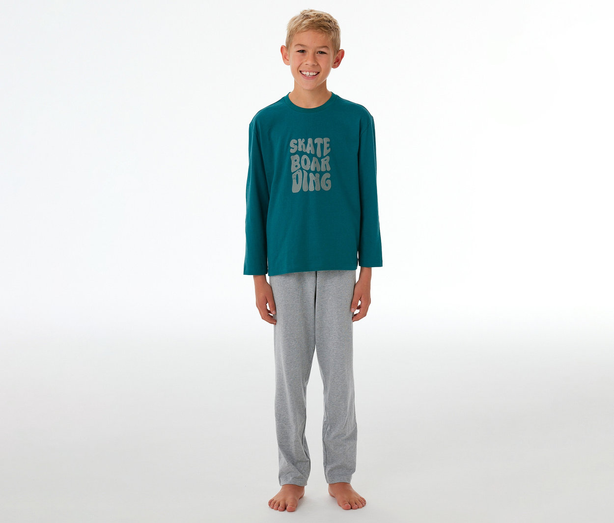 Garçon souriant portant un pyjama enfant avec un t-shirt à manches longues bleu pétrole, l'inscription grise "SKATE BOARDING" et un pantalon de pyjama gris.