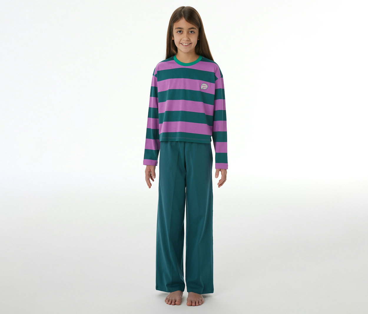 Jeune fille souriante en pyjama à manches longues rayé violet et vert, pantalon large vert, pieds nus.