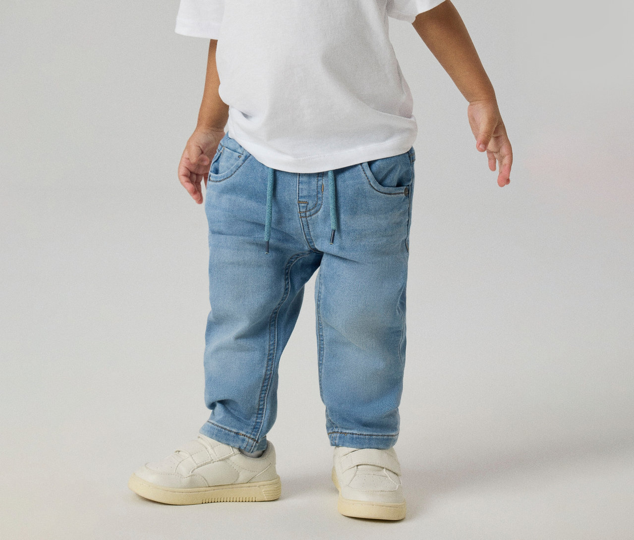 Enfant portant un t-shirt blanc, un jean NAME IT Slim Sweat bleu clair avec un cordon de serrage bleu et des baskets blanches.
