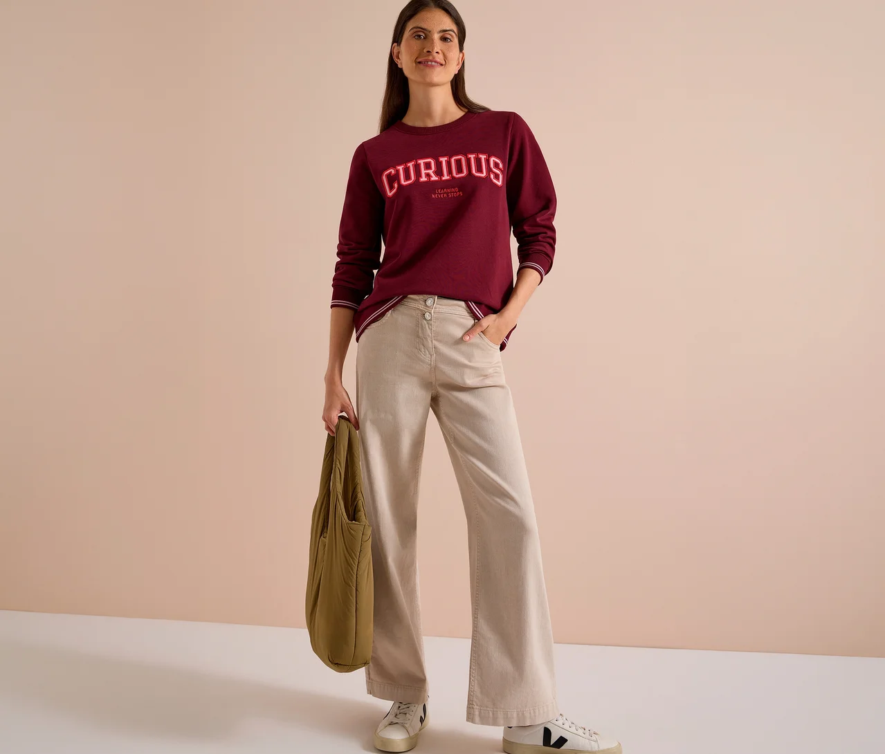 Femme souriante portant un sweatshirt CECIL bordeaux avec l'inscription « CURIOUS », un pantalon beige et des baskets blanches, tenant un sac vert olive.