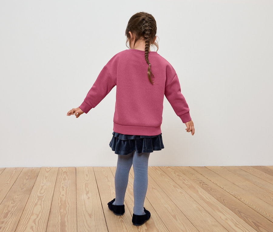 Une fille avec une tresse se tient dos à la caméra, portant un sweat-shirt rose, une jupe bleue et des collants gris.