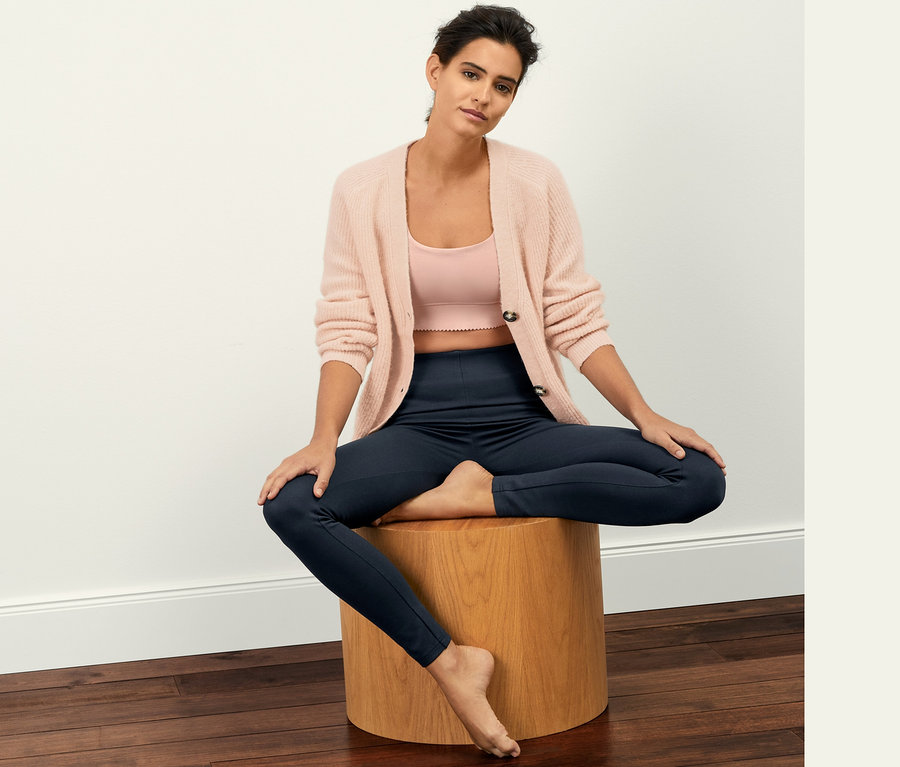Une femme est assise sur un tabouret en bois et porte un legging en jersey lourd, un débardeur rose et un cardigan rose.