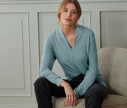 Une femme est assise sur une chaise et porte un t-shirt manches longues bleu fumée avec encolure en V.