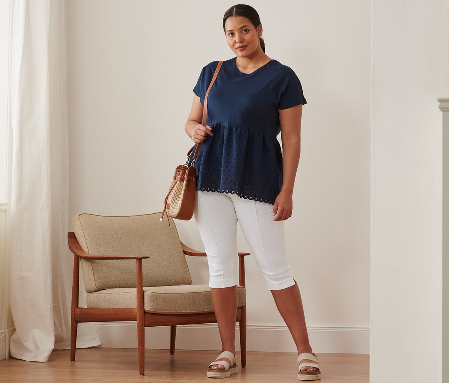 Une femme debout porte un t-shirt bimatière à broderie bleu foncé, un corsaire stretch, des sandales à plateau en cuir et un sac.