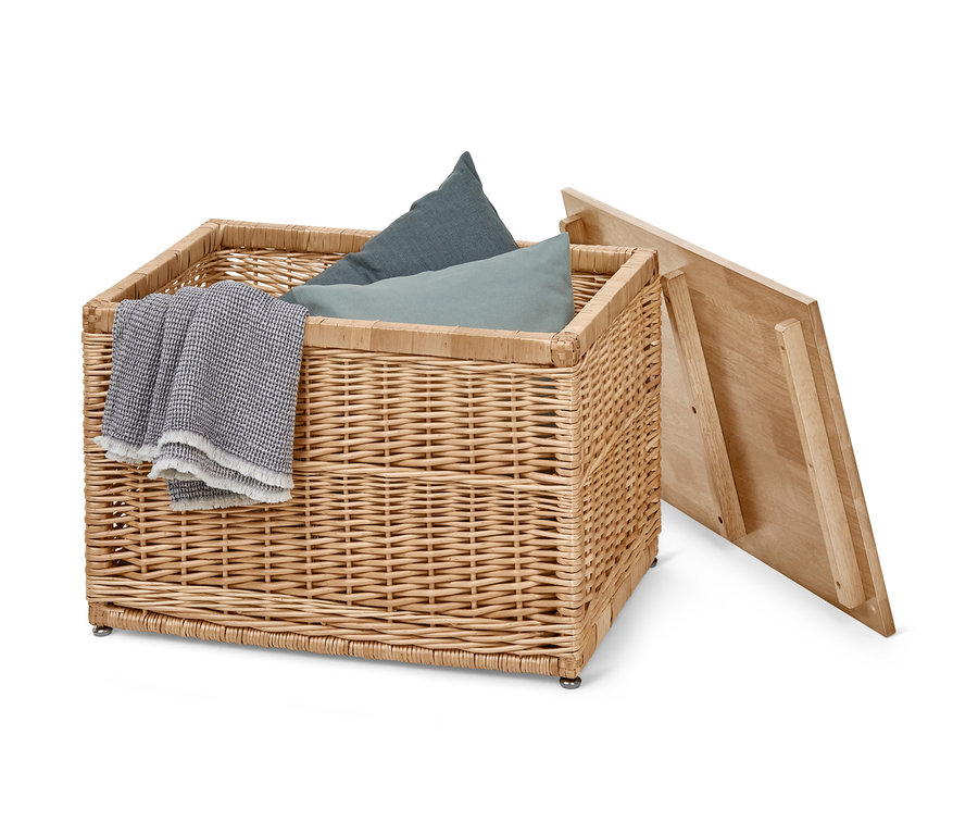 Un coffre tressé contenant des coussins bleus et une couverture grise, avec un couvercle en bois ouvert.