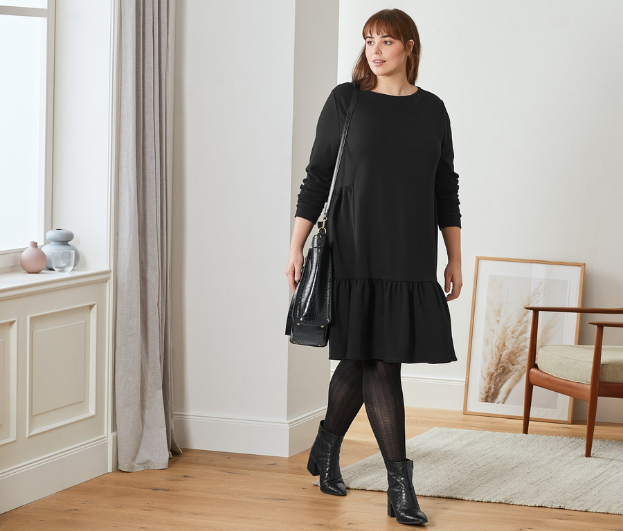 Une femme marche et porte une robe molletonnée noire à volant, des bottines en cuir avec relief et des collants à motif ajouré. Elle porte un sac de shopping noir sur l'épaule.