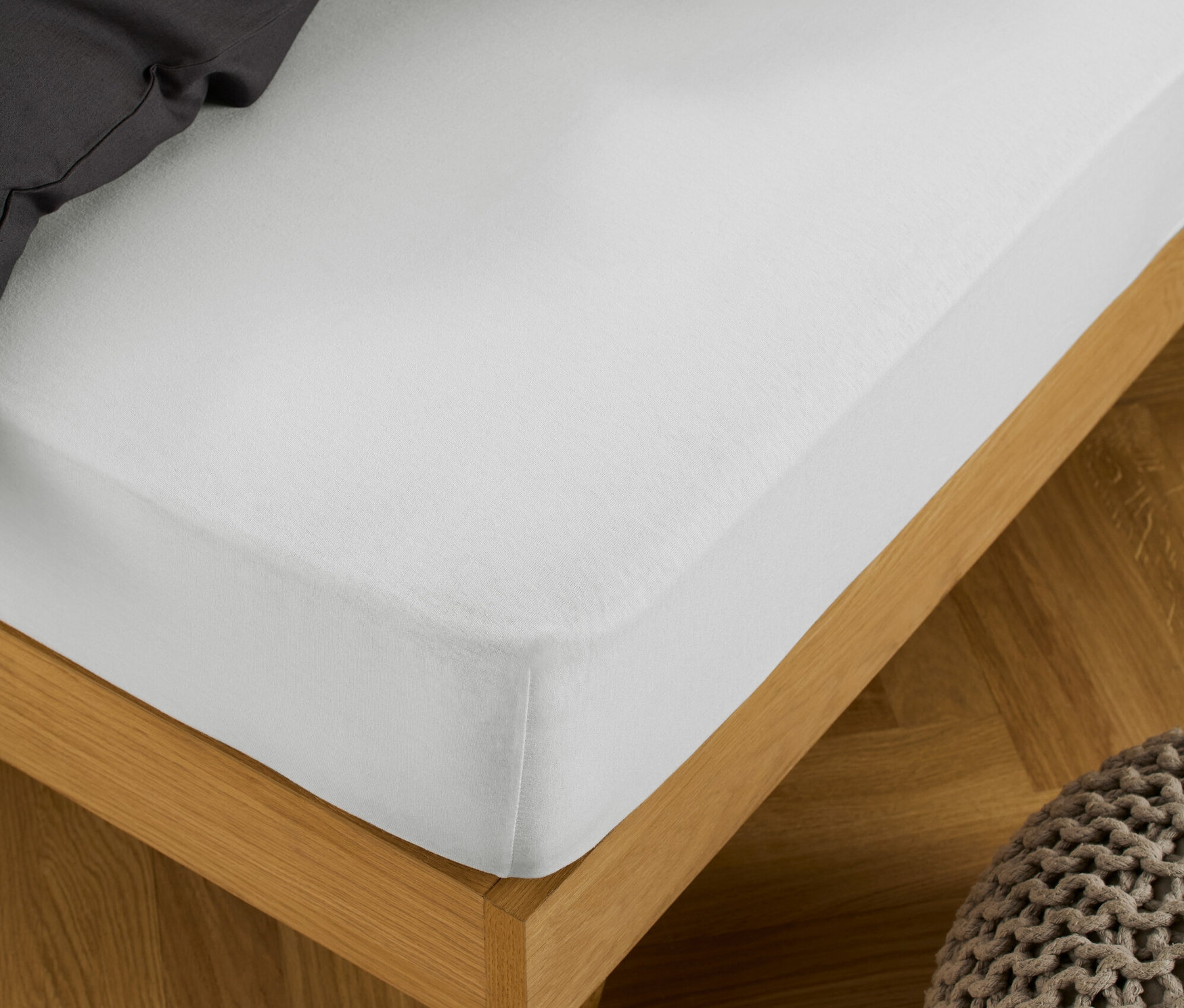 Un drap-housse en jersey blanc est étiré sur un lit avec un cadre en bois, à côté duquel se trouve un pouf tricoté.