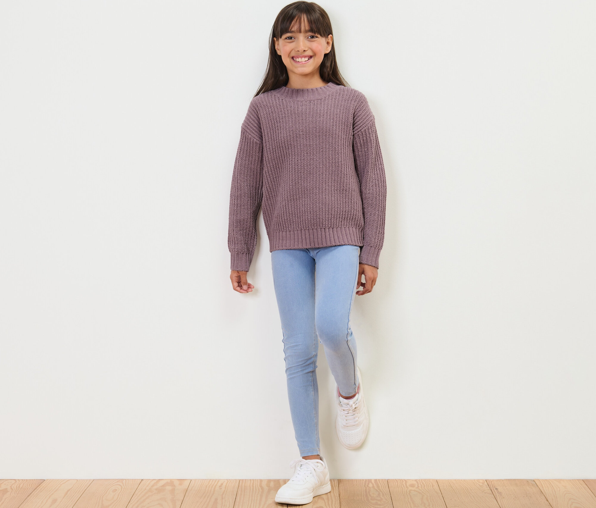 Une fille debout porte un pull en chenille mauve pour enfant, un jean clair et des baskets blanches.