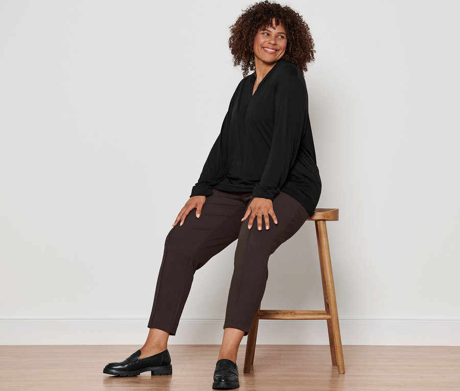 Une femme est assise sur un tabouret et porte un t-shirt manches longues avec encolure en V noir, un pantalon de jogging avec nervure marron foncé et des mocassins en cuir.