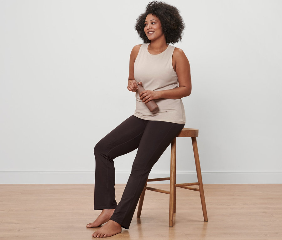 Femme assise sur une chaise en bois, portant un haut de sport beige et un pantalon de sport modelant chocolat, tenant une bouteille marron.