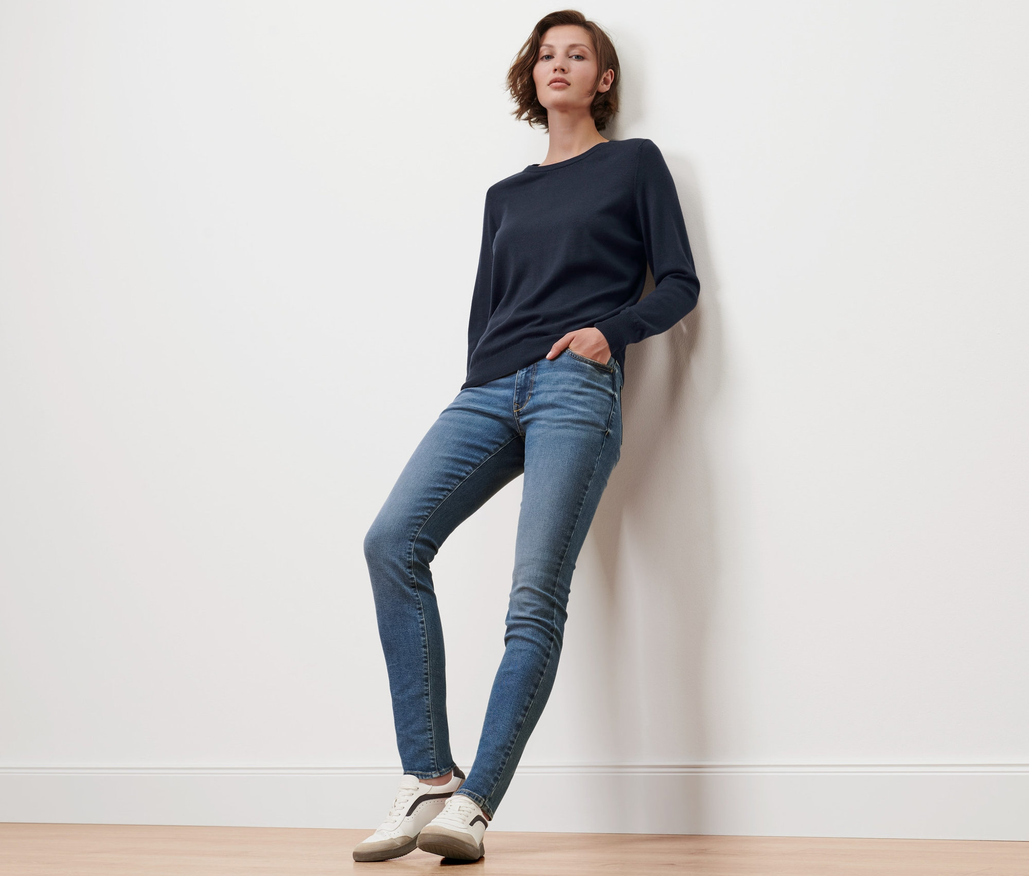 Femme appuyée contre un mur portant un pull en maille fine de mérinos bleu foncé.