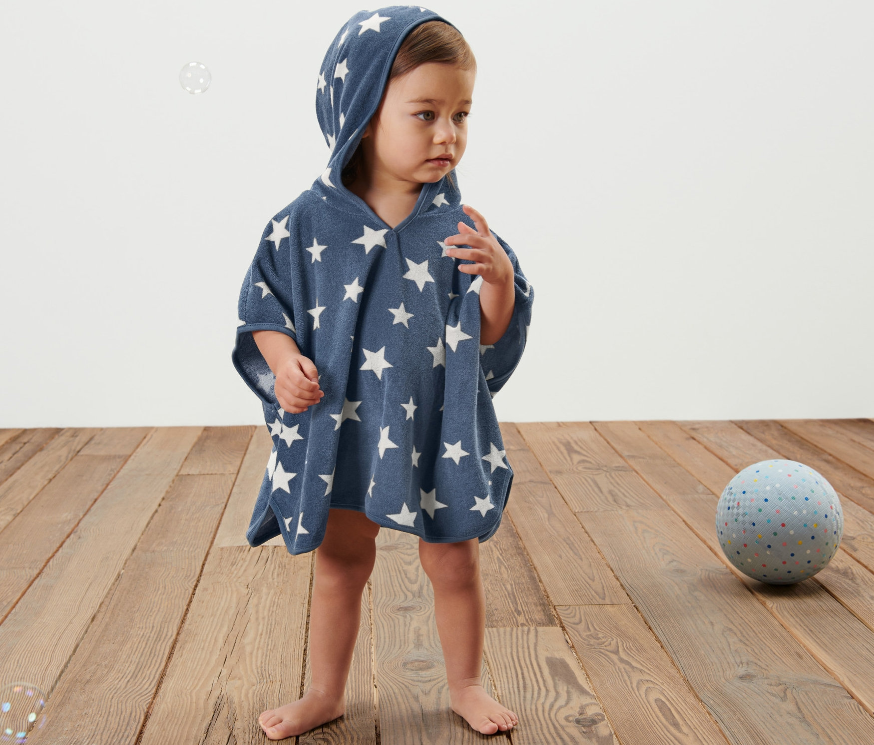 Un enfant se tient sur un plancher en bois et porte un poncho bleu en tissu éponge avec une capuche et des étoiles blanches.