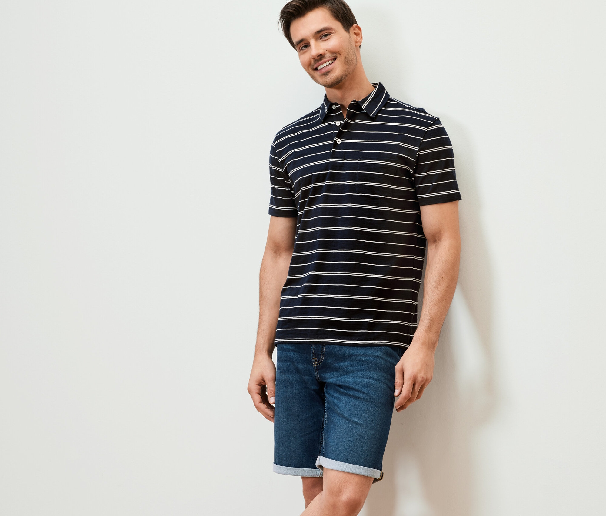 Un homme souriant appuyé contre un mur portant un polo en jersey bleu foncé à rayures et un short en jean.