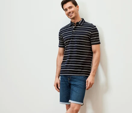 Un homme souriant appuyé contre un mur portant un polo en jersey bleu foncé à rayures et un short en jean.