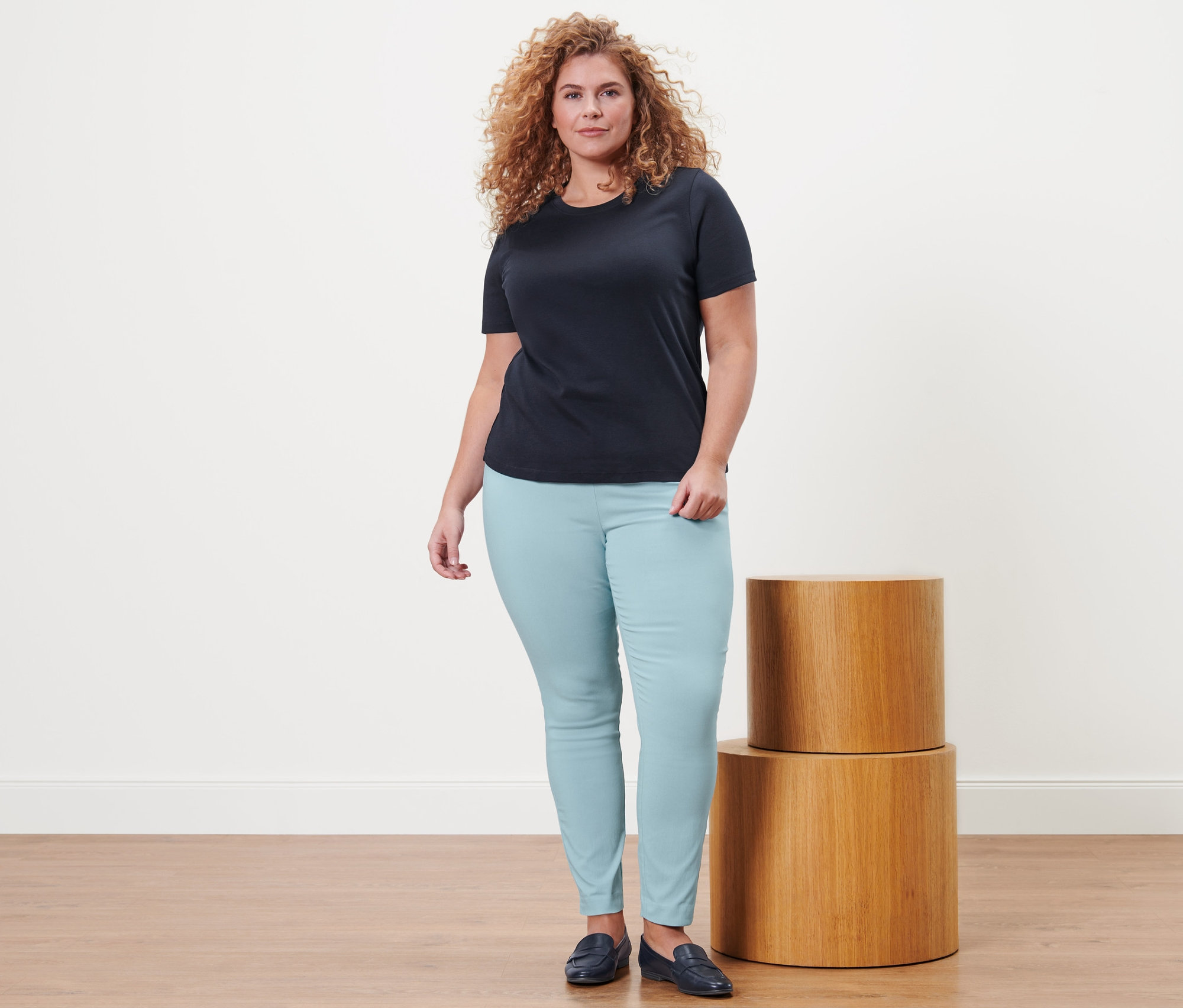 Une femme debout porte un t-shirt basique bleu foncé, un pantalon stretch et des loafers en cuir.