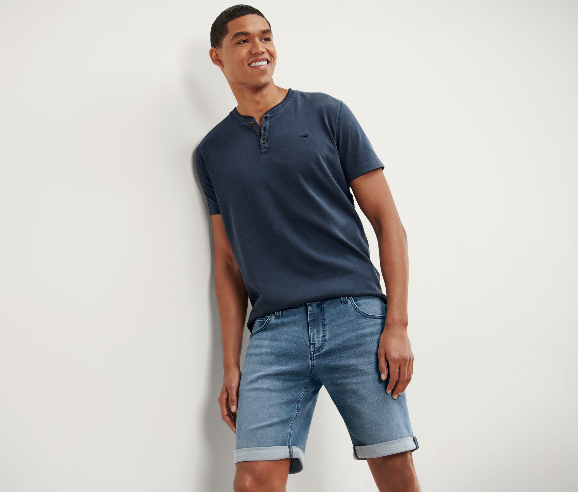 Un homme souriant, adossé à un mur, porte un t-shirt bleu et un short en jean « Mustang ».