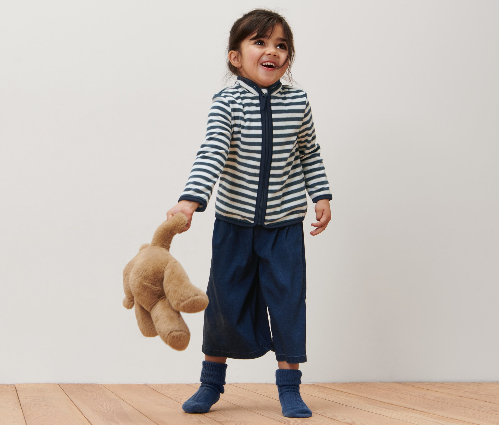 Fillette souriante tenant un ours en peluche et portant une jaquette en polaire marinière.