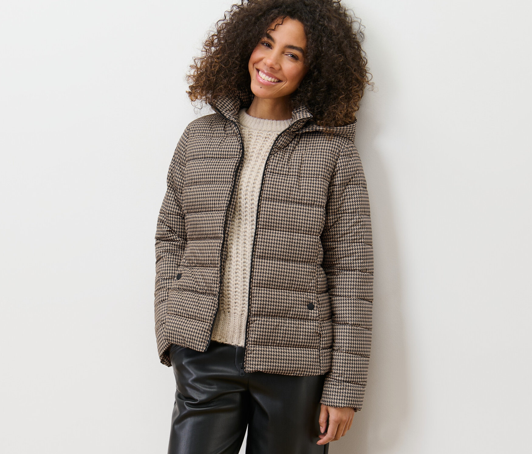 Une femme souriante s'appuie contre un mur et porte une veste matelassée imprimée avec capuche.