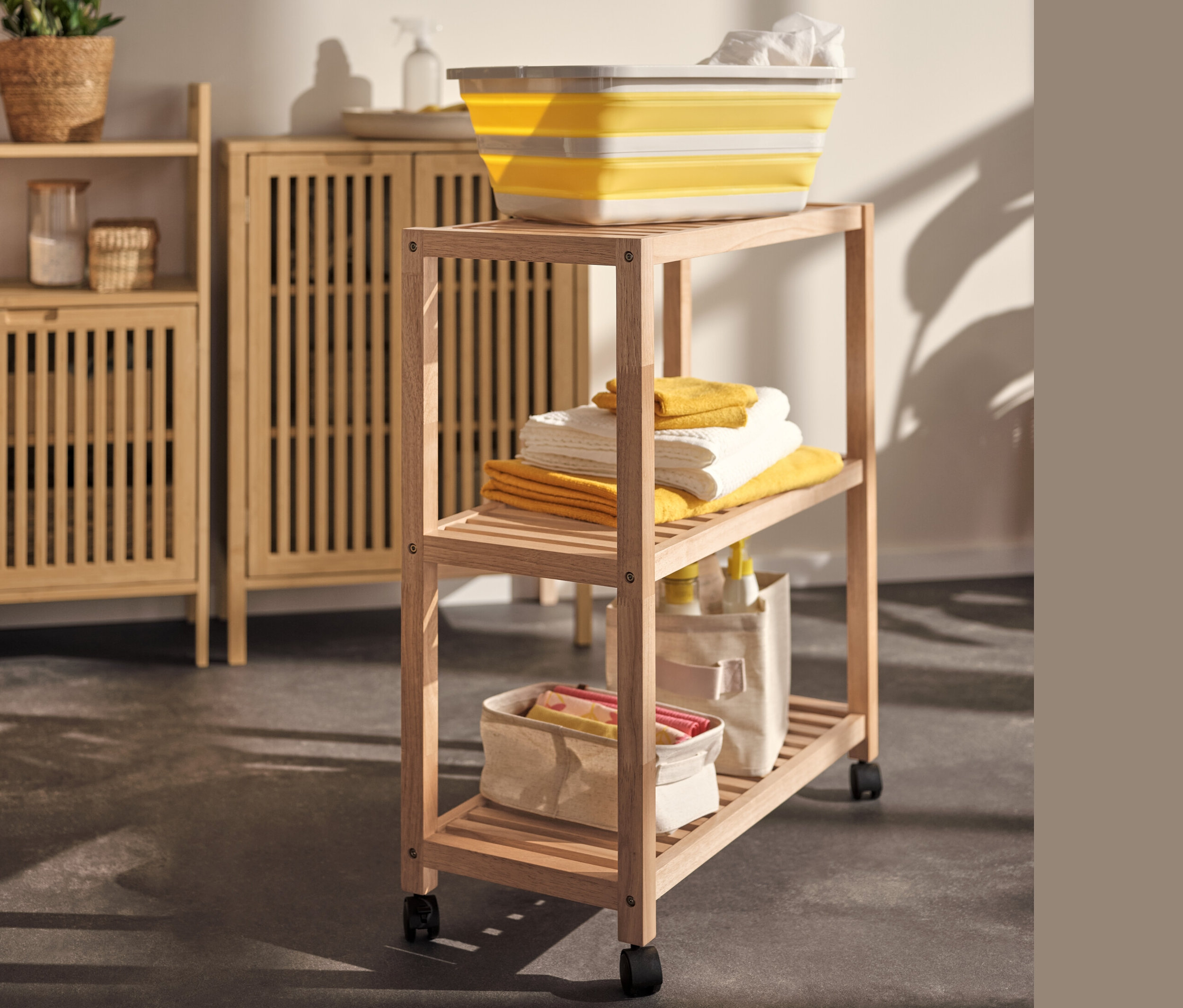 Panier à linge jaune pliable sur un étagère en bois massif à roulettes à côté d'un buffet.