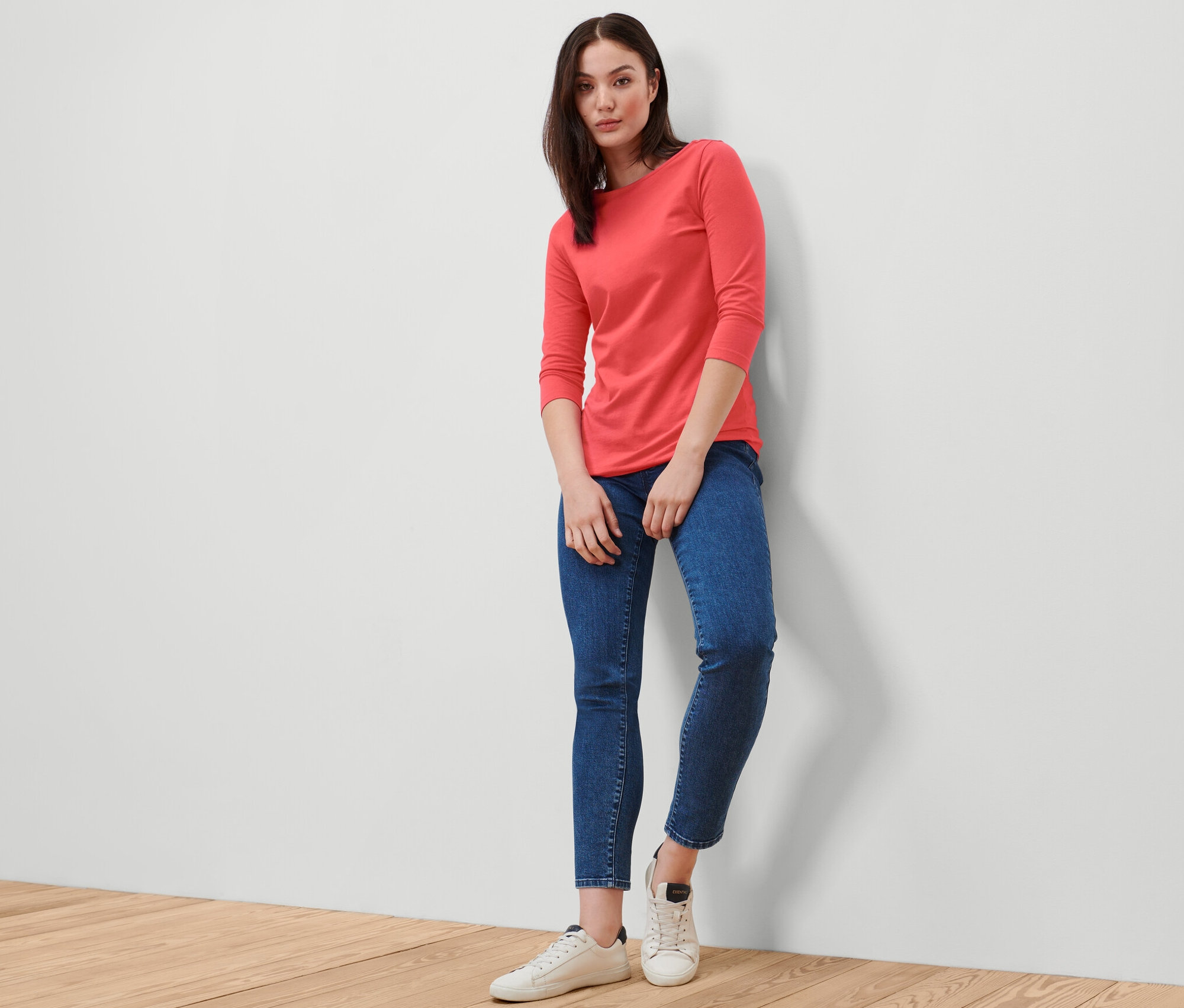 Femme posant en t-shirt corail manches 3/4, jean bleu et baskets blanches.