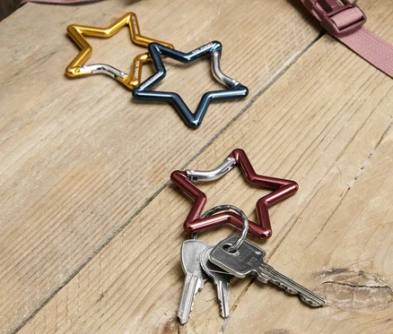 Trois mousquetons en forme d'étoile dorés, bleus et rouges sont posés sur une table en bois, avec des clés accrochées au mousqueton rouge. Un morceau de sac à dos est visible en arrière-plan.