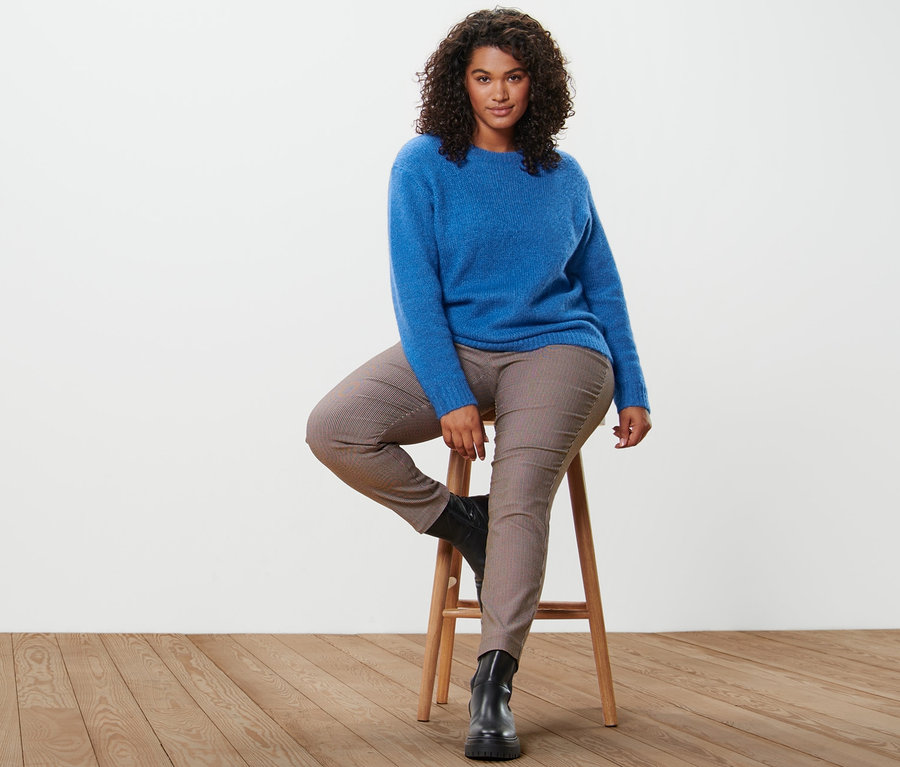Une femme est assise sur un tabouret et porte un pull bleu cobalt en grosse maille avec de la laine et un pantalon stretch à carreaux.