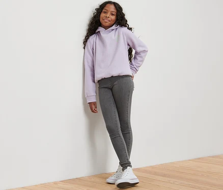 Une fille adossée au mur porte un tregging pour enfant – Fit « Jule », gris et un sweat à capuche violet.