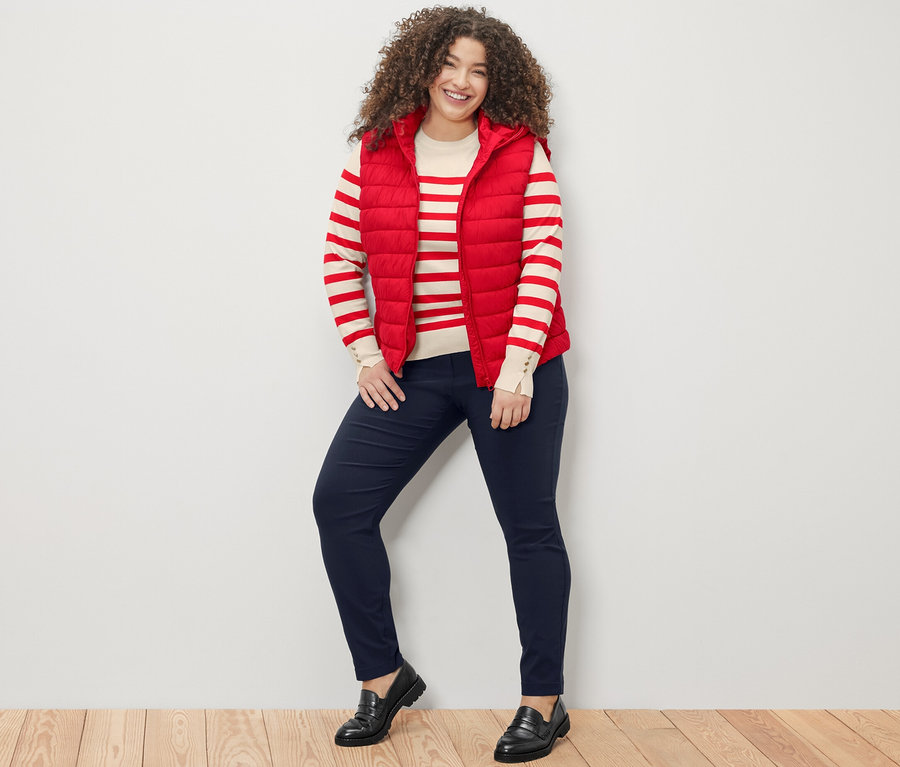 Une femme debout souriant à la caméra, portant un pull en maille fine rayé rouge, un gilet sans manches matelassé à capuche rouge et un pantalon en bengaline bleu foncé.