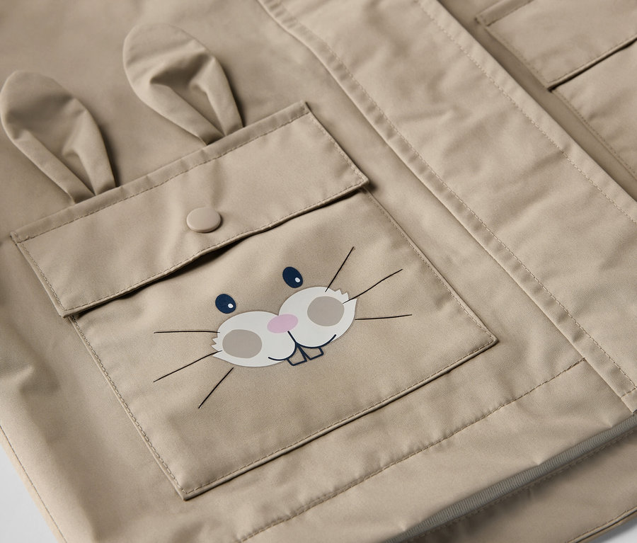 Gros plan sur une veste de pluie marron pour enfant avec une poche ornée d'un visage de lapin.