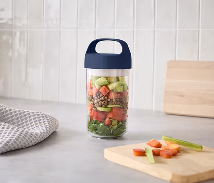 Un verre avec des couches de salade est posé sur un plan de travail de cuisine à côté d'une planche à découper avec des légumes coupés.