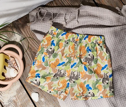 Un boardshort à motif animalier posé sur une table en bois à côté d'un jouet de sable et de coquillages.