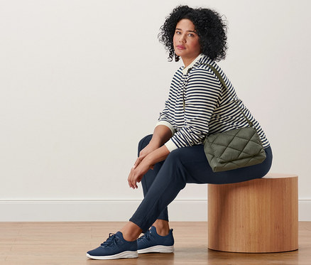 Femme assise sur un tabouret en bois, portant un sweat-shirt à rayures, un pantalon de jogging avec nervure et des baskets en cuir. Un sac matelassé à bandoulière croisé sur son corps.