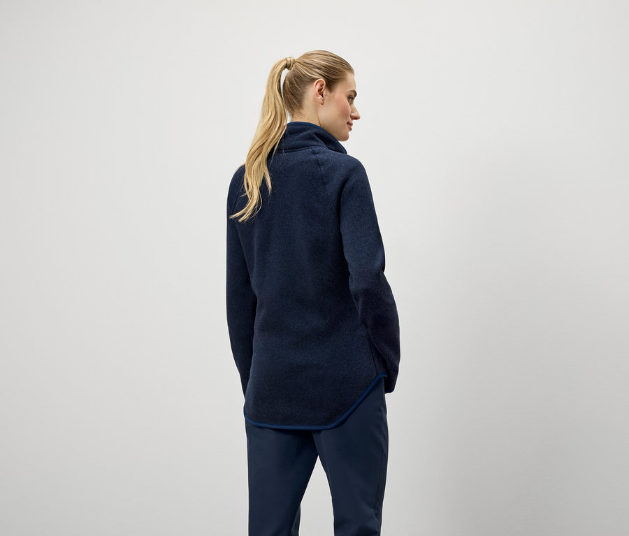 Vue de dos, une femme aux cheveux blonds attachés en queue de cheval, portant un pull bleu marine et un pantalon.