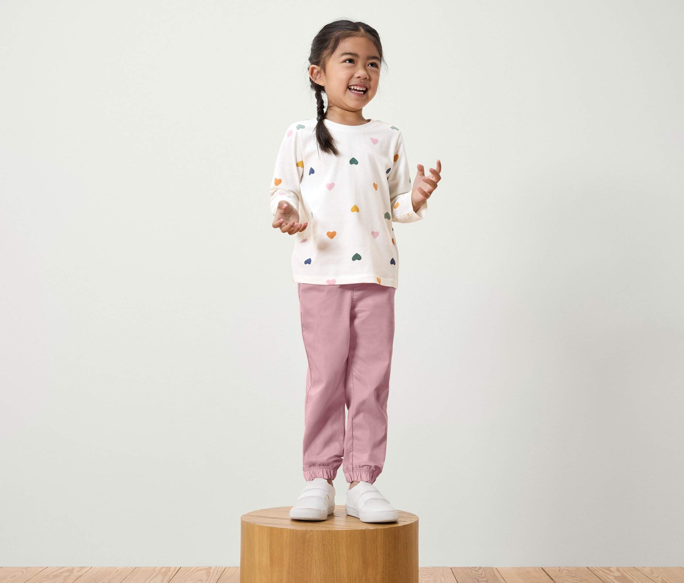 Une fillette avec une tresse se tient sur un socle en bois. Elle porte un t-shirt blanc à manches longues avec des cœurs colorés et un pantalon rose.