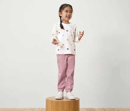 Une fillette avec une tresse se tient sur un socle en bois. Elle porte un t-shirt blanc à manches longues avec des cœurs colorés et un pantalon rose.