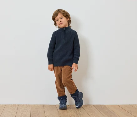 Un garçon se tient devant un mur blanc. Il porte un pull bleu foncé, un pantalon marron et des chaussures bleues.