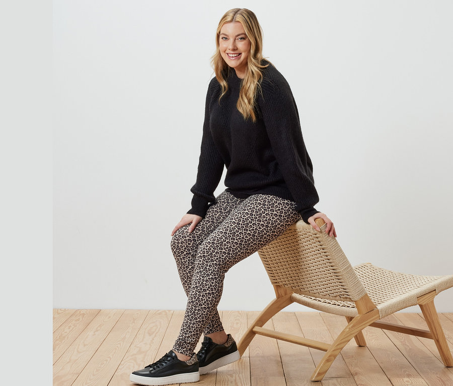 Une femme est assise sur un fauteuil en osier et porte un pull en maille noir et un legging imprimé.