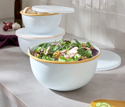 Trois bols blancs avec un bord coloré sont empilés les uns sur les autres, le plus grand bol contient de la salade, à côté se trouve une planche à découper jaune avec du concombre coupé.