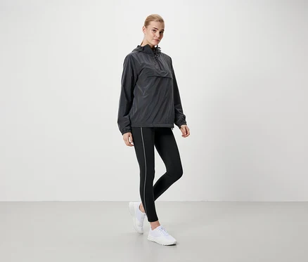 Une femme blonde se tient debout, portant une veste à capuche grise, un legging noir et des chaussures blanches.
