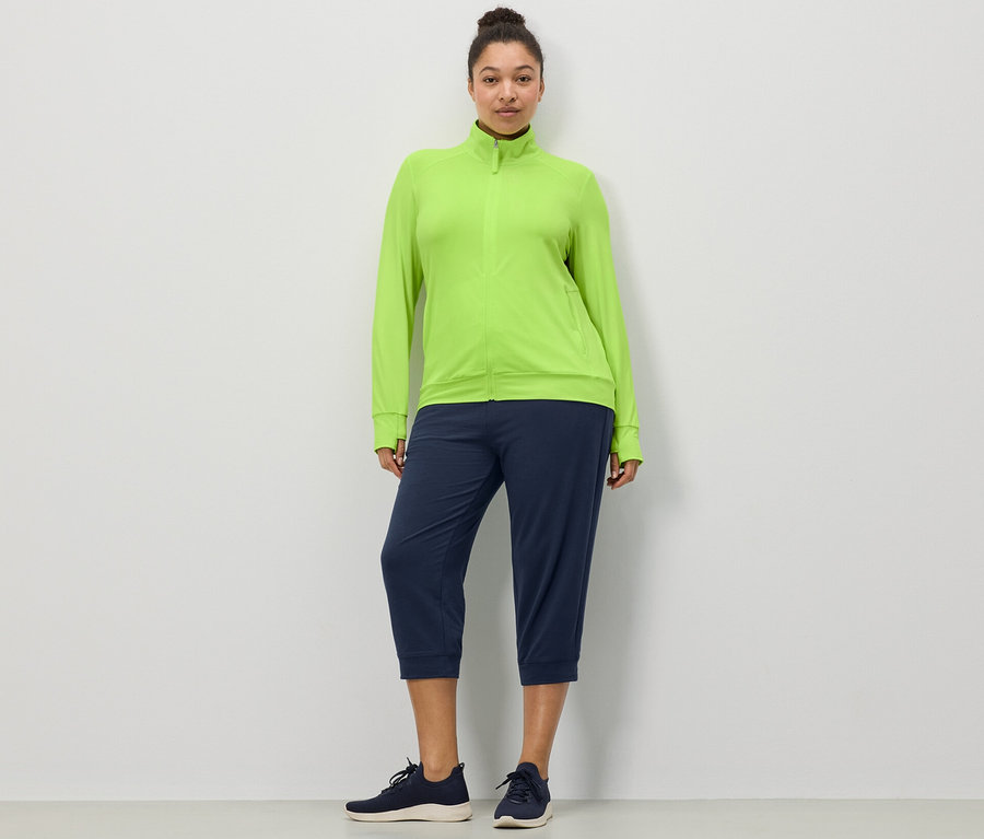 Une femme debout devant un mur blanc porte un pantalon sweat 3/4 bleu foncé et une veste fonctionnelle vert citron.