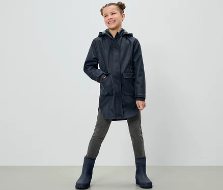 Une jeune fille souriante pose dans un manteau softshell pour enfants, un pantalon gris et des bottes bleues.