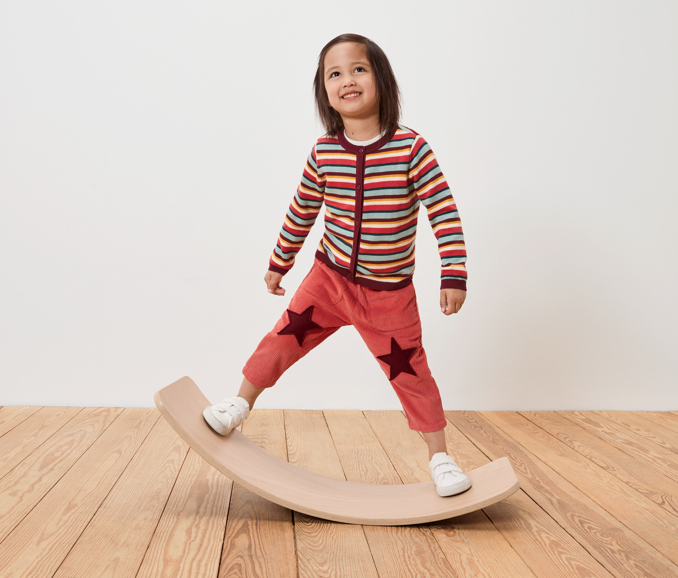 Un enfant se tient sur une planche d'équilibre en bois, portant un pull rayé et un pantalon avec des étoiles.