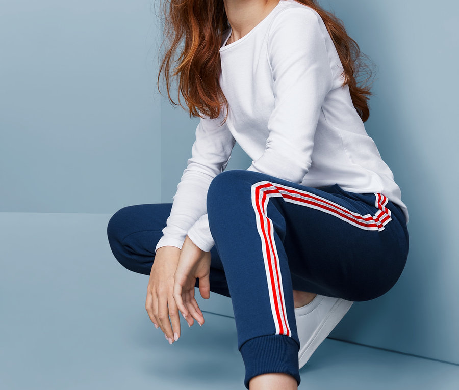 Une fille portant un pantalon de survêtement avec des rayures rouges et blanches, un t-shirt blanc et des chaussures blanches est accroupie sur un fond bleu.